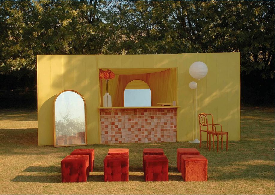 An outdoor set in a field — a soft yellow bar, arched mirror, and red velvet stools